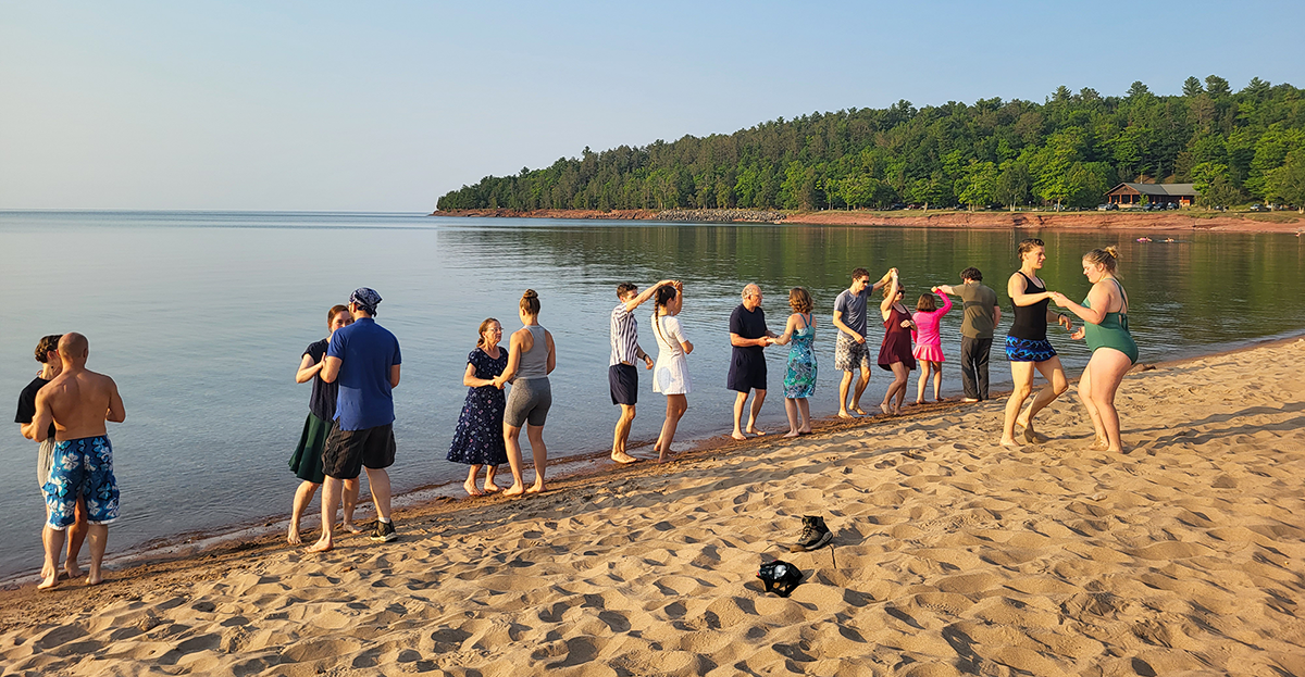 people dancing bachata on a sandy beach with a blue sky and green trees in the background